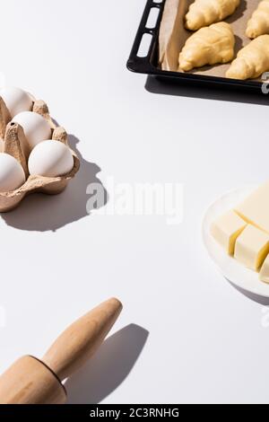 selective focus of raw croissants on baking tray near rolling pin, butter, eggs on white background Stock Photo