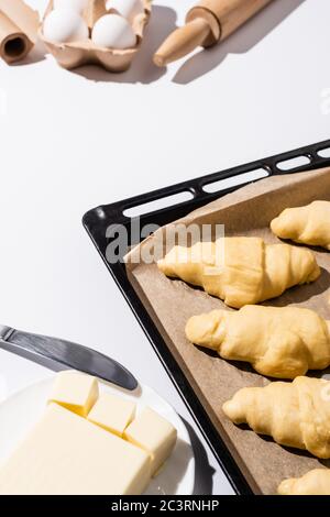 selective focus of raw croissants on baking tray near rolling pin, parchment paper, butter, eggs, knife on white background Stock Photo