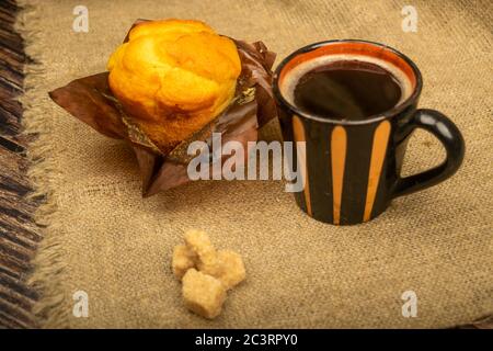 A fresh cupcake, a mug of coffee, and chunks of brown cane sugar on a rough-textured homespun cloth. Close up Stock Photo
