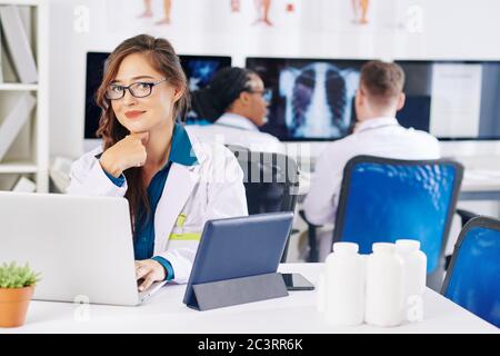 Portrait of pretty smiling young female doctor in glasses sitting at office desk with laptop, tablet computer and containers with medicine, her collea Stock Photo