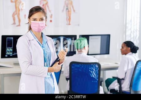 Young female doctor in medical mask holding tablet computer and looking at camera, her colleagues discussing x-rays and medical tests of patients in b Stock Photo