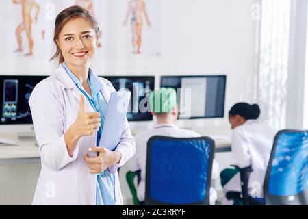 Portrait of pretty young general practitioner with folder in hands showing thumbs-up and smiling at camera Stock Photo