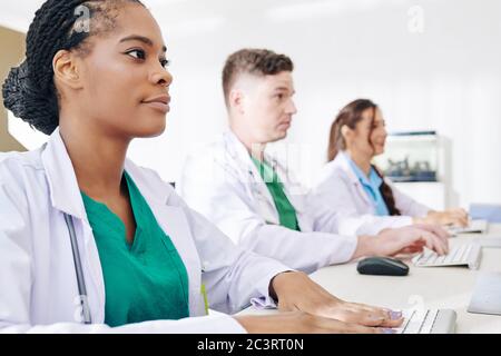Young Black general practitioner filling form on computer after vising patient, her colleagues working on computers nearby Stock Photo