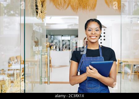 Pretty happy small coffeeshop owner standing at entrance doors with tablet computer in hands ready to invite first customers Stock Photo