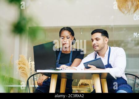 Serious young bakery owners sitting at small table, discussing financial issues, paying bills and ordering products online Stock Photo