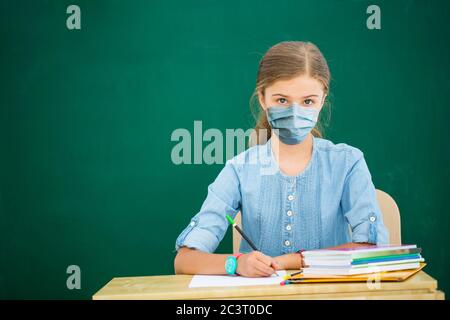 Back to school  after quarantine girl sitting in the classroom against blackboard. Child in school with book and accessories. Education concept with c Stock Photo