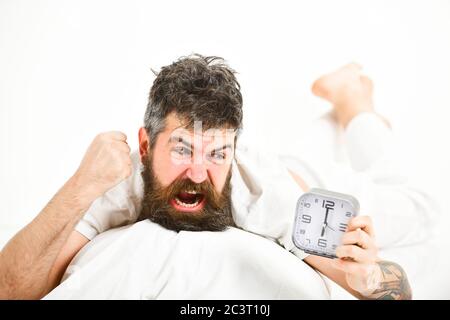 Hipster with beard and mustache awake because of alarm clock. Morning wake up concept. Man suffers in morning, wants to destroy alarm clock, white background. Guy with furious face holds alarm clock. Stock Photo