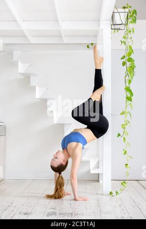 Young woman doing a handstand stretch yoga pose using a wooden frame for support as she balances on her hands in a high key gym with green plants in a Stock Photo