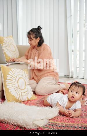 Young woman sitting on the floor and working on laptop when little daughter crawling near by Stock Photo