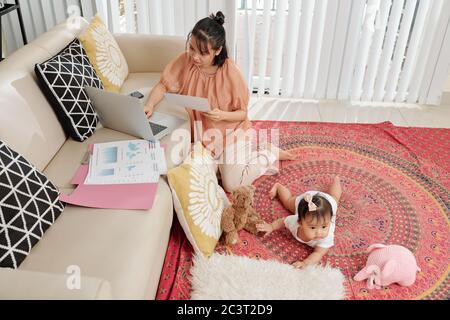 Young Asian businesswoman working with financial documents when staying at home with little baby girl Stock Photo