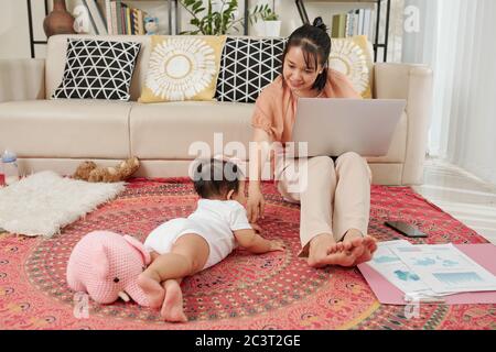 Young smiling businesswoman sitting on the floor with laptop and playing with little baby girl when working from home during quarantine Stock Photo