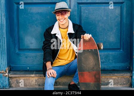 Cheerful and happy teenager sit down outside home with blue old door in background - city urban lifestyle and trendy generation z people concept - you Stock Photo