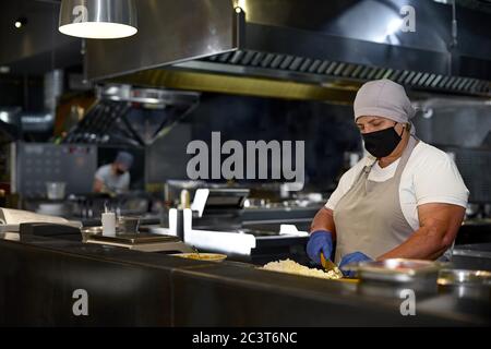 cooking, profession and people concept - woman in age chef wearing protective black mask on her face while working in restaurant kitchen Stock Photo