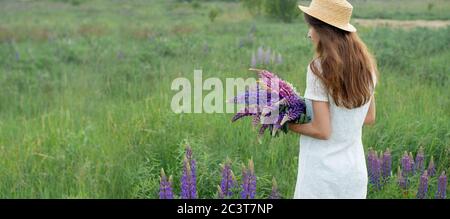 Beautiful woman in lupine field banner. Cute girl holding a bouquet of lupines in white dress and hat. Summer time. Back view Stock Photo