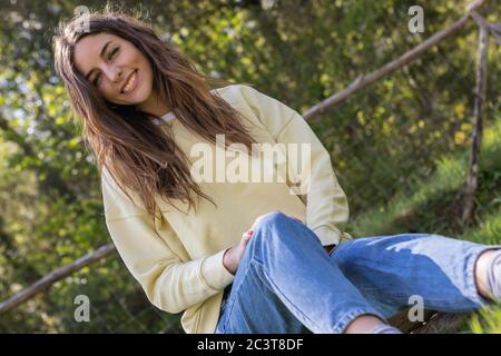 Pretty young brunette woman smiling and looking at camera sitting in a park outdoors on a sunny day. She is wearing blue jeans and a yellow sweater Stock Photo