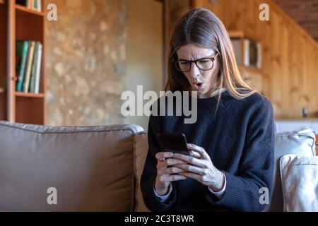 Shocked pretty young brunette woman with glasses typing on the smartphone sitting on the sofa at home wearing black sweater Stock Photo
