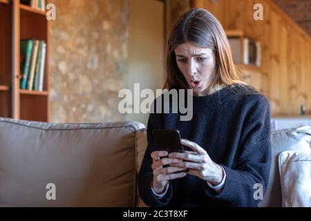 Shocked pretty young brunette woman typing on the smartphone sitting on the sofa at home wearing black sweater Stock Photo