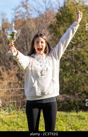 Excited pretty young woman holds a trophy outdoors in a park. She is wearing a light sweatshirt and has long brown hair. Stock Photo
