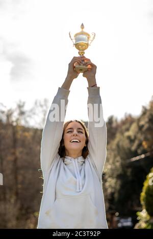 Pretty smiling young woman holds a trophy outdoors in a park. She is wearing a light sweatshirt and has long brown hair. Stock Photo