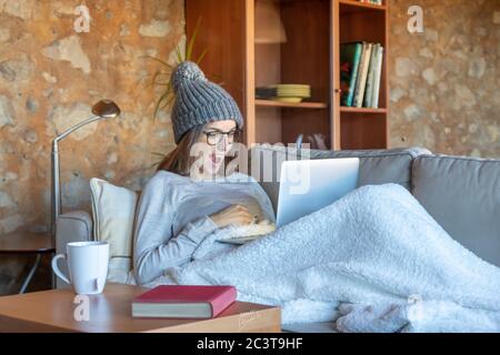 Surprised pretty young woman sitting on the sofa at home looking at the laptop screen. She is wearing a wool hat and glasses. Horizontal image.