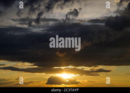 Fiery orange tropical sunset in a cloudy sky over open countryside with the sun shining out between layers of clouds low on the horizon Stock Photo