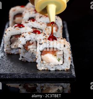 Delicious sushi roll served on black slate. In the frame, the hand of a chef pouring food with sweet sauce.