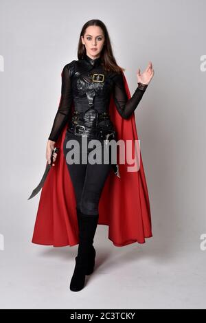 Portrait of brunette girl wearing black leather catsuit & red cloak.  full length standing pose, isolated against a studio background. Stock Photo