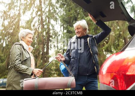 Low angle portrait of modern senior couple opening trunk of car and smiling happily while going on hike in forest Stock Photo