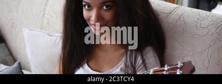 Dark-skinned girl sitting on sofa playing ukulele Stock Photo