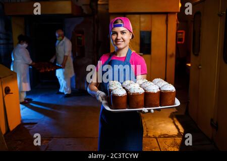 Girl baker holds a tray with hot pastries in the bakery. She is wearing a denim frock and a cap. Production of bakery products. Rack with fresh crispy Stock Photo