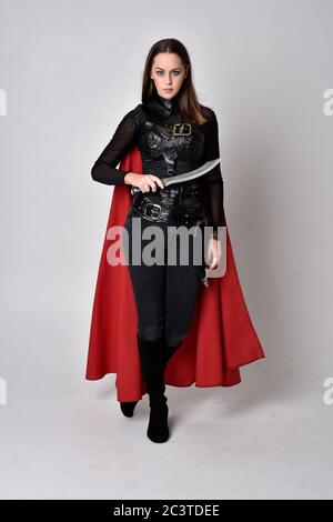 Portrait of brunette girl wearing black leather catsuit & red cloak.  full length standing pose, isolated against a studio background. Stock Photo