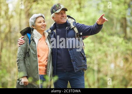 Waist up portrait of modern senior couple wearing activewear enjoying hike in forest, focus on smiling man pointing away, copy space Stock Photo