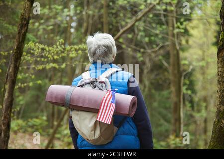 Back view at active senior man wearing backpack with American flag while enjoying hike in forest, copy space Stock Photo