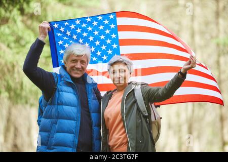 Waist up portrait of active senior couple holding American flag and smiling at camera while enjoying hike in forest, copy space Stock Photo