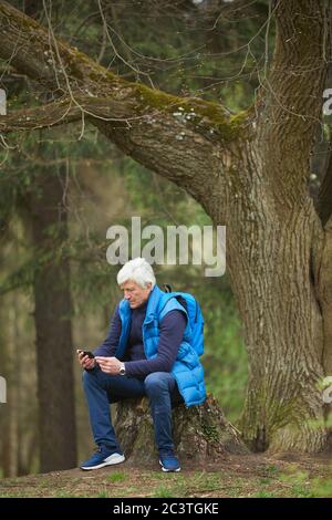 Vertical full length portrait of active senior man using smartphone while resting during hike in autumn forest, copy space above Stock Photo