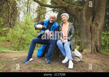 Full length portrait of cheerful senior couple enjoying hot tea from flask while resting during hike in autumn forest, copy space Stock Photo