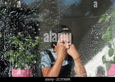 Young teenage boy, 12-13, looking bored through a freshly washed window , dueing Covid 19 lockdown, Surrey, UK Stock Photo