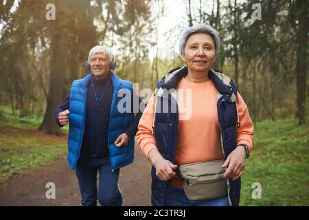 Waist up portrait of active senior couple running towards camera in beautiful autumn forest and smiling happily Stock Photo