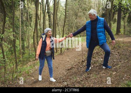 Full length portrait of active senior couple helping each other while climbing on hill during hike in forest, copy space Stock Photo