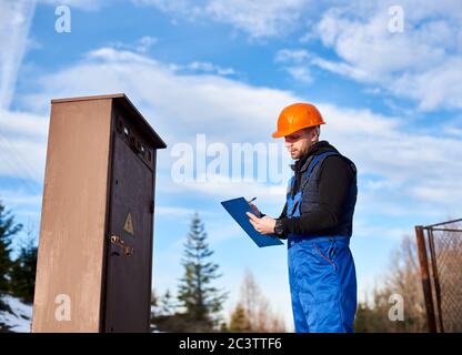 Oil inspector in protective overalls and orange helmet making notes next to a transformer outdoors against blue sky with white clouds. Concept of petroleum industry. Stock Photo