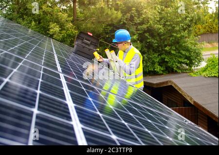 Photovoltaic business. Solar panels installation and commissioning. Renewable green energy in a private home. Worker with electric tools on the roof. Stock Photo