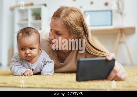 Warm toned portrait of mature mother taking selfie photo with cute mixed race baby while playing on carpet at home, copy space Stock Photo