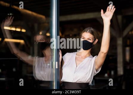 Portrait of waitress with face mask standing at the door in restaurant, waving.