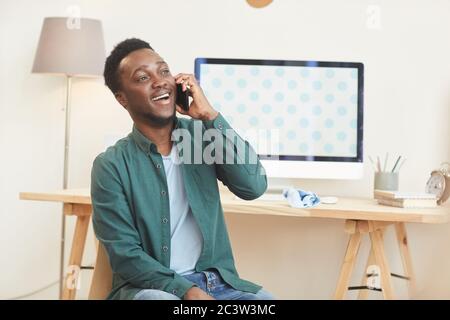 Portrait of young African-American man speaking by phone and smiling while sitting at desk in modern home interior, copy space Stock Photo