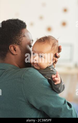 Vertical back view portrait of African-American man holding cute mixed-race baby looking at camera while posing in cozy home interior Stock Photo