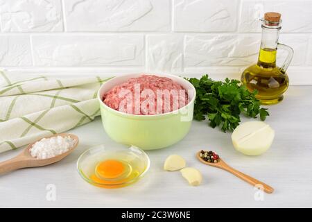 Fresh minced meat in a green bowl, egg, onion, garlic, salt and pepper on a kithcen table. Raw ingredients for cutlets or meatballs. Cook at home. Stock Photo