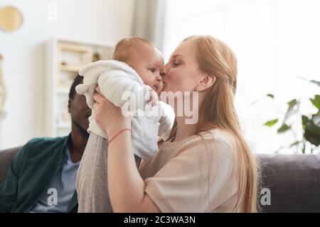 Portrait of happy interracial family at home, focus on Caucasian woman kissing cute baby in foreground, copy space Stock Photo