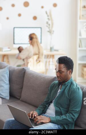 Vertical portrait of modern interracial family working from home, focus on African-American man using laptop in foreground, copy space Stock Photo