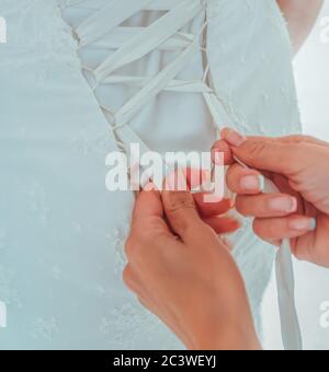 Morning preparation of the bride. Closeup of bridesmaid tying bow on brides wedding dress. White ribbon bow on the back of wedding dress. Bride in Stock Photo