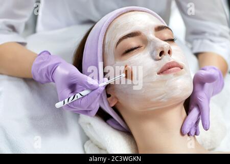 beautician applies the mask to the face of beautiful young woman in the spa salon. Anti-aging, facial skin care concept. Stock Photo
